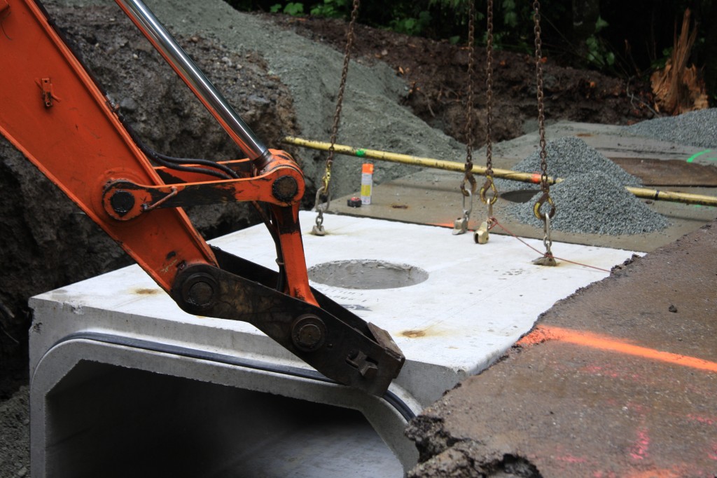 Maneuvering culvert sections into place using crane and excavator.